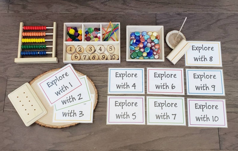 Puede incluir: Un conjunto de materiales de aprendizaje Montessori para contar y explorar los n&uacute;meros. El conjunto incluye un &aacute;baco de madera, una bandeja con fichas de n&uacute;meros, una bandeja con gemas de vidrio de colores, una tabla de clavijas de madera y tarjetas con las palabras "Explorar con" y los n&uacute;meros del 1 al 10.
