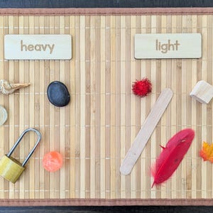 May include: A wooden educational toy with the words "heavy" and "light" on separate wooden blocks. Various objects are arranged on a bamboo mat to demonstrate weight differences, including a shell, stone, lock, and feather.