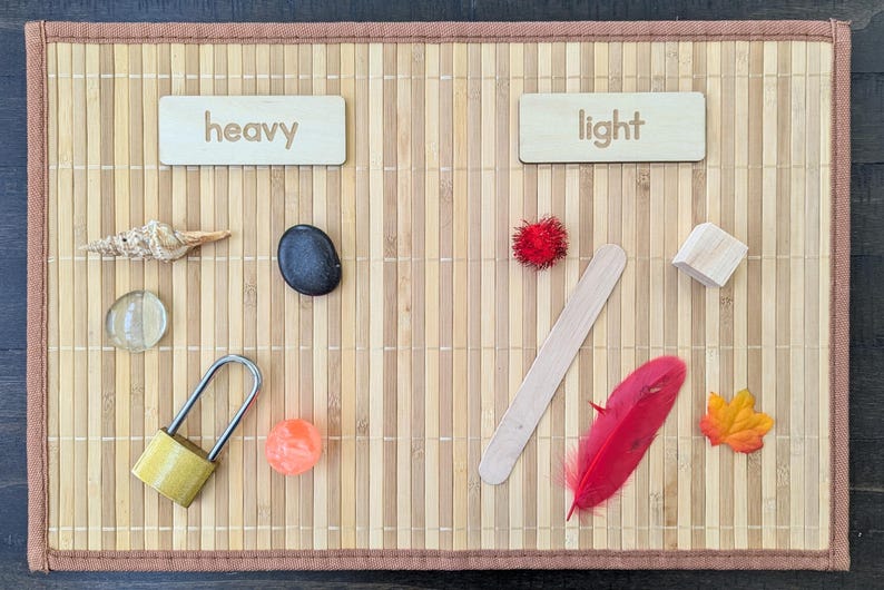 May include: A wooden educational toy with the words "heavy" and "light" on separate wooden blocks. Various objects are arranged on a bamboo mat to demonstrate weight differences, including a shell, stone, lock, and feather.