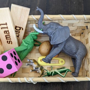 May include: A woven basket filled with educational toys. Includes wooden blocks with "small" and "big" text, a pink die, a gray elephant, green grasshoppers, a brown egg, a yellow safety pin, and a small animal figurine.