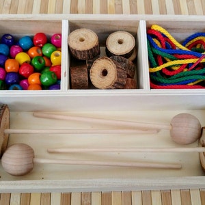Op de afbeelding: Houten doos met vakken die kleurrijke houten kralen, houten pinnen, houten ringen en gekleurd touw bevatten. De doos is open en de inhoud is zichtbaar.