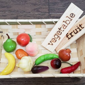 May include: A woven basket filled with toy fruits and vegetables. The basket has two wooden signs, one that says "vegetable" and one that says "fruit".