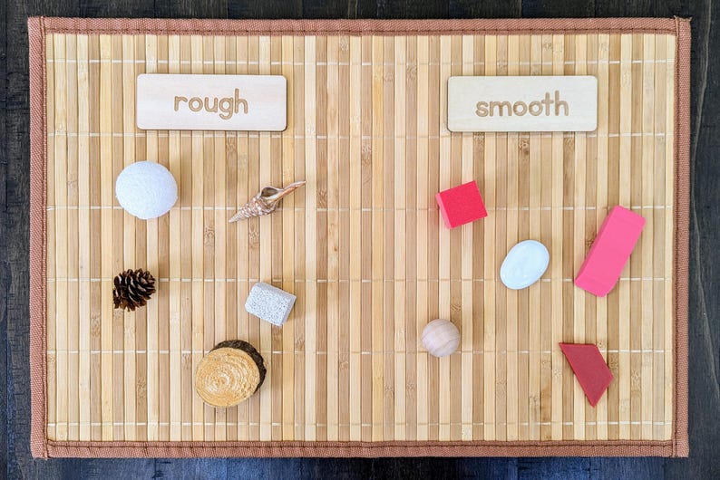 May include: A sensory learning activity with objects labeled "rough" and "smooth". The "rough" side includes a white ball, pinecone, shell, stone, and wood slice. The "smooth" side has red and pink shapes, and a small ball.