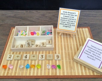 May include: A wooden tray with compartments filled with colourful beads and gems. There are also wooden blocks with numbers 1, 5, 7, 8, and 9.  Two cards with maths problems are on the table. The first card says "Cindy dropped 2 jewels in the lake, left 4 jewels in her backpack, and lost 3 jewels on the way to school. How many jewels did Cindy lose altogether?" The second card says "Tammy had 5 flowers and she gave 3 flowers to her friends. How many flowers does Tammy have left?"