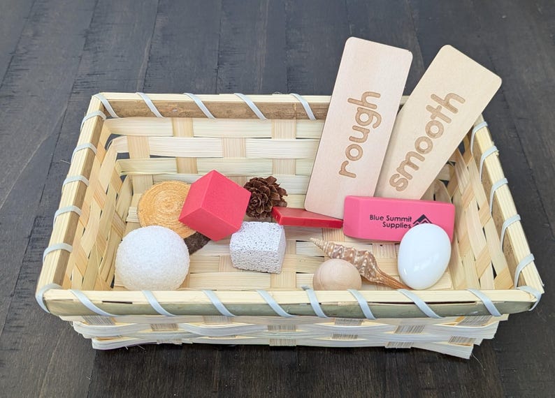 May include: A woven basket filled with various textured objects. Includes wooden cards labeled "rough" and "smooth", a red cube, a pine cone, a white sphere, a piece of pumice, and a pink rectangular object.