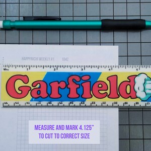 May include: A white sheet of paper with a grid pattern and the text "HAPPY NICHI WEEKLY #1 1042" printed on it. A ruler with the word "Garfield" printed on it is placed on top of the paper. The ruler is yellow, blue, and red with a white cartoon character on it. A white label with the text "MEASURE AND MARK 4.125" TO CUT TO CORRECT SIZE" is placed on the paper.