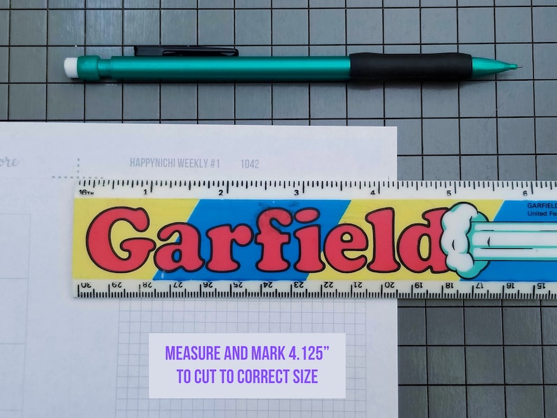 May include: A white sheet of paper with a grid pattern and the text "HAPPY NICHI WEEKLY #1 1042" printed on it. A ruler with the word "Garfield" printed on it is placed on top of the paper. The ruler is yellow, blue, and red with a white cartoon character on it. A white label with the text "MEASURE AND MARK 4.125" TO CUT TO CORRECT SIZE" is placed on the paper.
