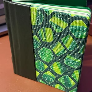 May include: A hardcover journal with a green and black color scheme. The cover features a pattern of green dice on a dark green background. The spine is black, and the pages have green edges. The journal is open, revealing the lined pages.