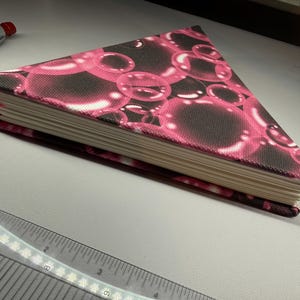 Puede incluir: Un cuaderno triangular con una cubierta con estampado de burbujas rosas y negras. El cuaderno tiene un marcador de cinta rosa. Las páginas son de color crema. Un bolígrafo rojo es visible en el fondo.