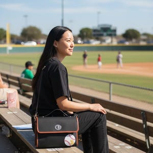 Può includere: Borsa a tracolla nera con finiture marroni e toppa da baseball. La borsa ha una chiusura argentata e una lunga tracolla. La persona indossa una maglietta nera, jeans neri e scarpe da ginnastica bianche, seduta su una panchina di legno in un campo da baseball.