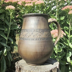 May include: A brown ceramic pitcher with a handle and decorative carvings. The pitcher is sitting on a tree stump in front of a green leafy background.