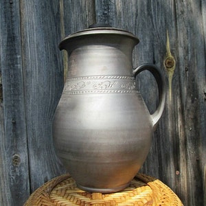May include: A large, gray ceramic pitcher with a lid and a handle. The pitcher has a decorative design around the middle.