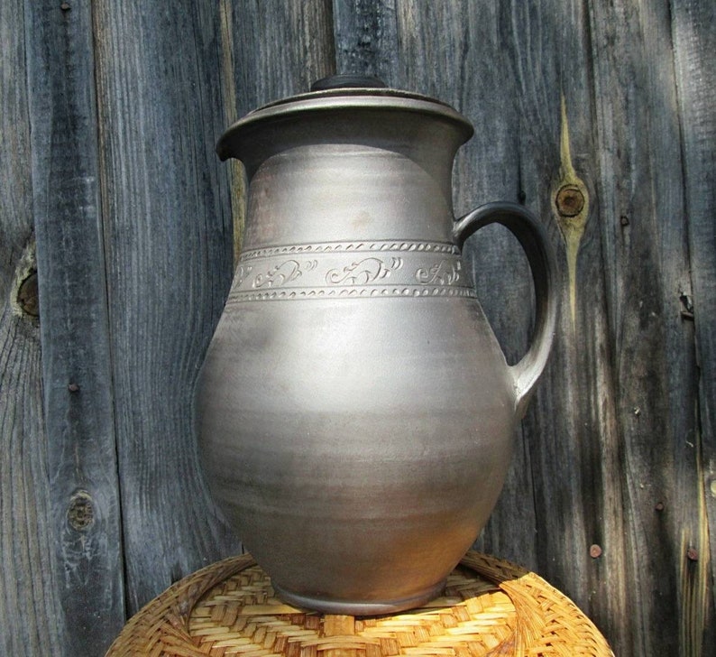 May include: A large, gray ceramic pitcher with a lid and a handle. The pitcher has a decorative design around the middle.