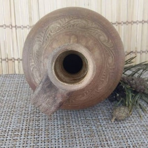 May include: A brown clay jug with a wide mouth and a handle. The jug has a decorative design carved into the surface.