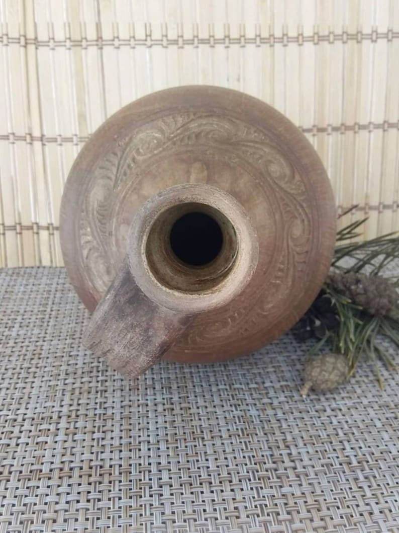 May include: A brown clay jug with a wide mouth and a handle. The jug has a decorative design carved into the surface.