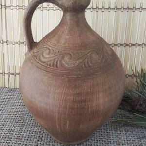 May include: A brown clay jug with a carved design and a handle. The jug is sitting on a woven mat.
