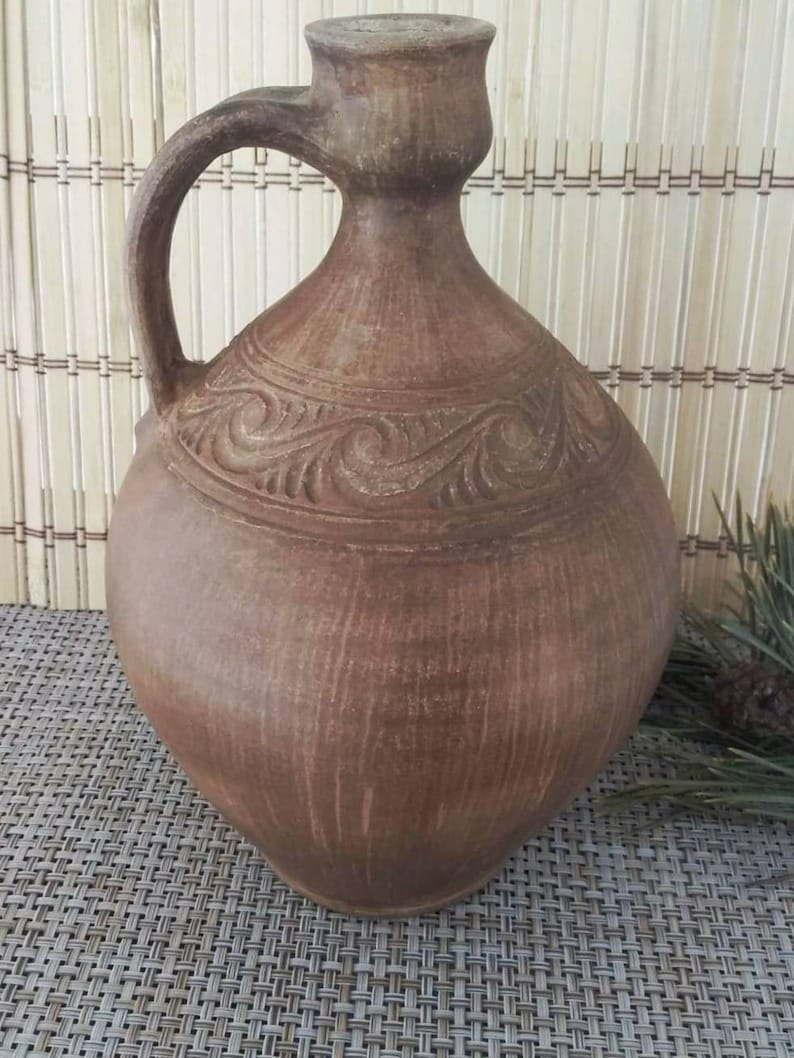 May include: A brown clay jug with a carved design and a handle. The jug is sitting on a woven mat.