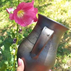 May include: A dark brown ceramic pitcher with a handle and decorative carvings. The pitcher is being held by a hand in front of a pink flower.