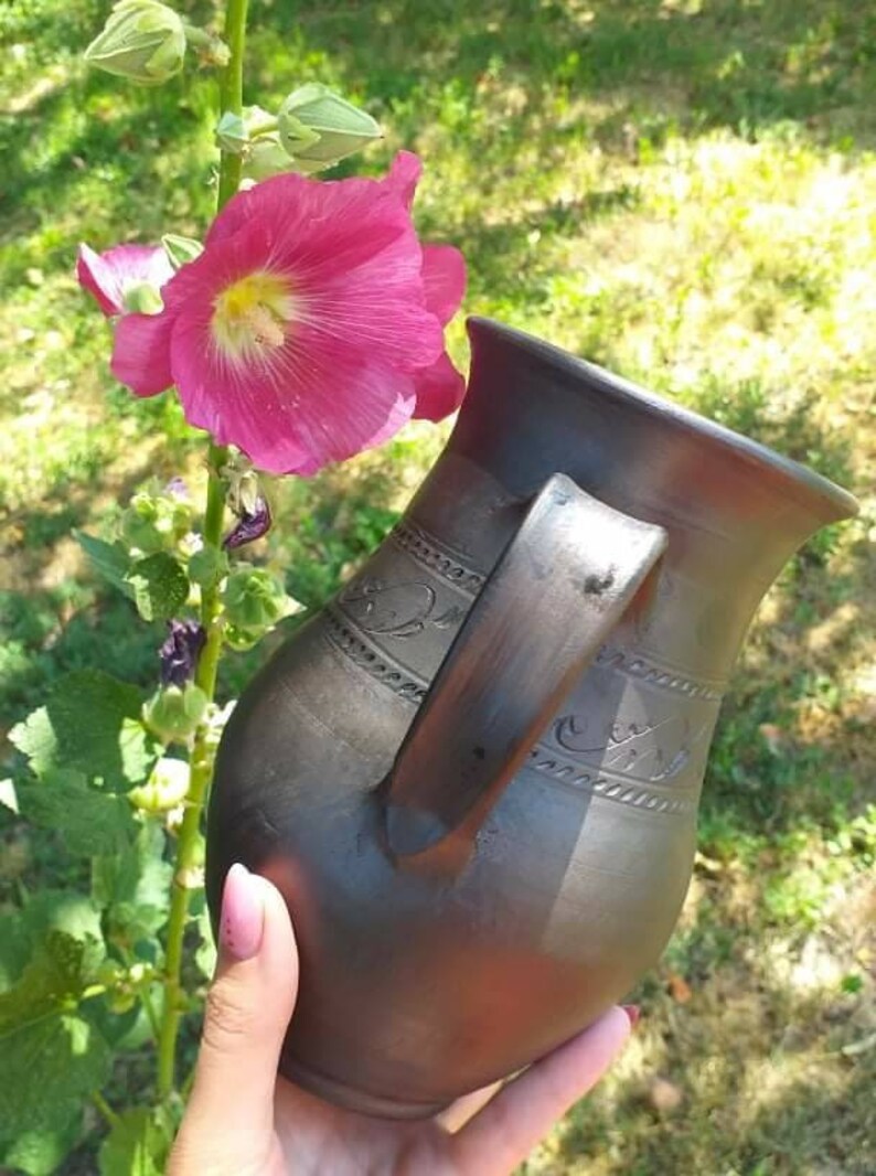 May include: A dark brown ceramic pitcher with a handle and decorative carvings. The pitcher is being held by a hand in front of a pink flower.