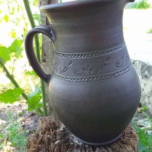 May include: A dark brown ceramic pitcher with a handle and a decorative design around the middle. The pitcher is sitting on a weathered wooden stump.