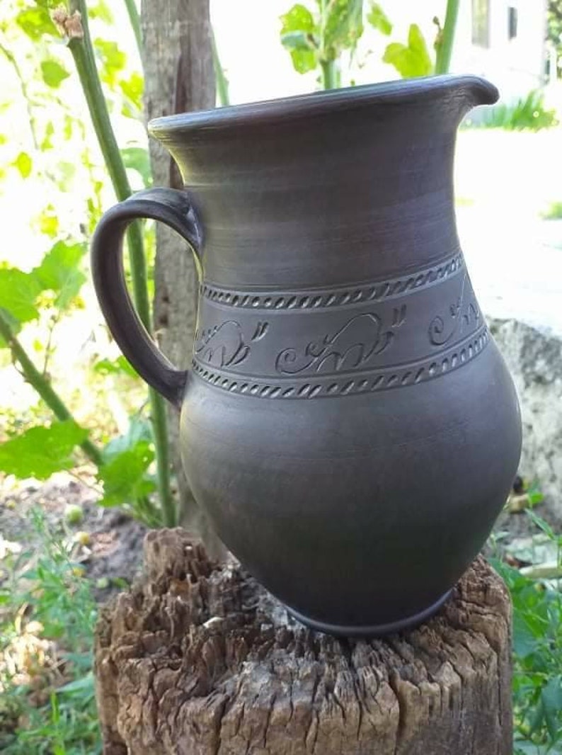 May include: A dark brown ceramic pitcher with a handle and a decorative design around the middle. The pitcher is sitting on a weathered wooden stump.