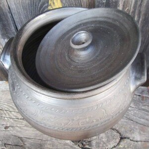 May include: A black ceramic pot with a lid, featuring a decorative design around the rim. The pot has a round shape and a small knob on the lid.