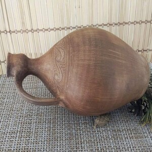 May include: A brown clay jug with a handle and a carved design around the middle. The jug is sitting on a woven mat.
