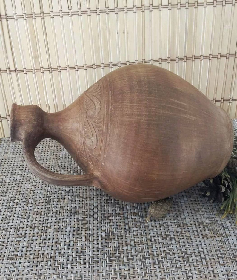 May include: A brown clay jug with a handle and a carved design around the middle. The jug is sitting on a woven mat.