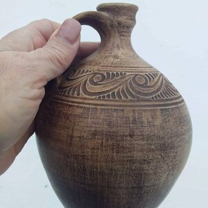 May include: A brown ceramic pitcher with a carved design. The pitcher has a wide base and a narrow neck with a handle.