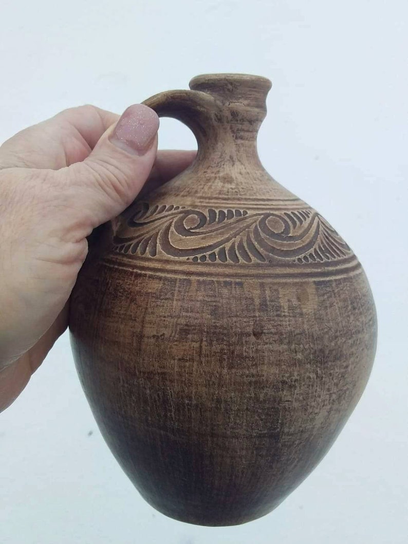 May include: A brown ceramic pitcher with a carved design. The pitcher has a wide base and a narrow neck with a handle.