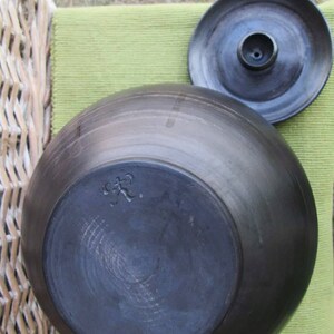 May include: Black ceramic pot with a lid. The pot has a round shape and a smooth surface. The lid has a small hole in the center. The bottom of the pot has a mark that says "C.P."