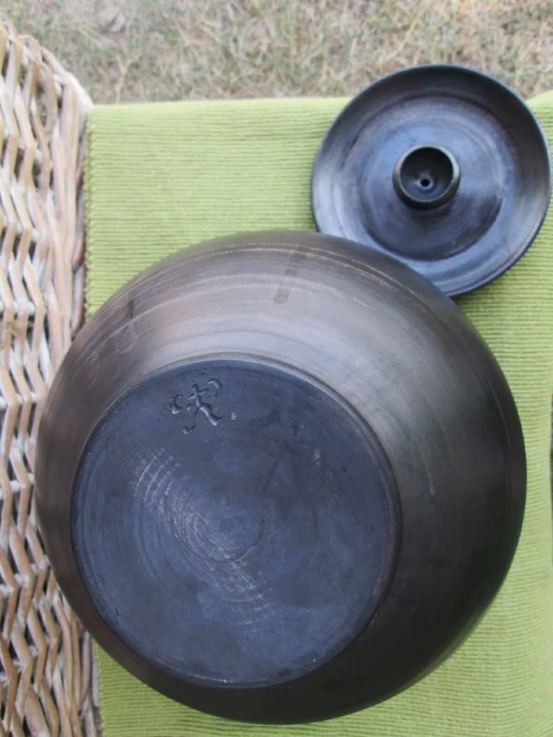 May include: Black ceramic pot with a lid. The pot has a round shape and a smooth surface. The lid has a small hole in the center. The bottom of the pot has a mark that says "C.P."