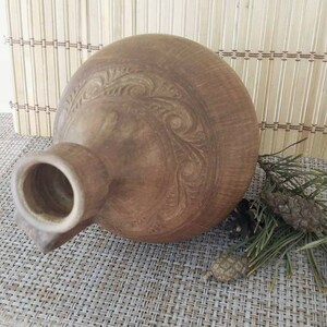 May include: A brown ceramic pitcher with a carved design. The pitcher has a wide mouth and a narrow neck.