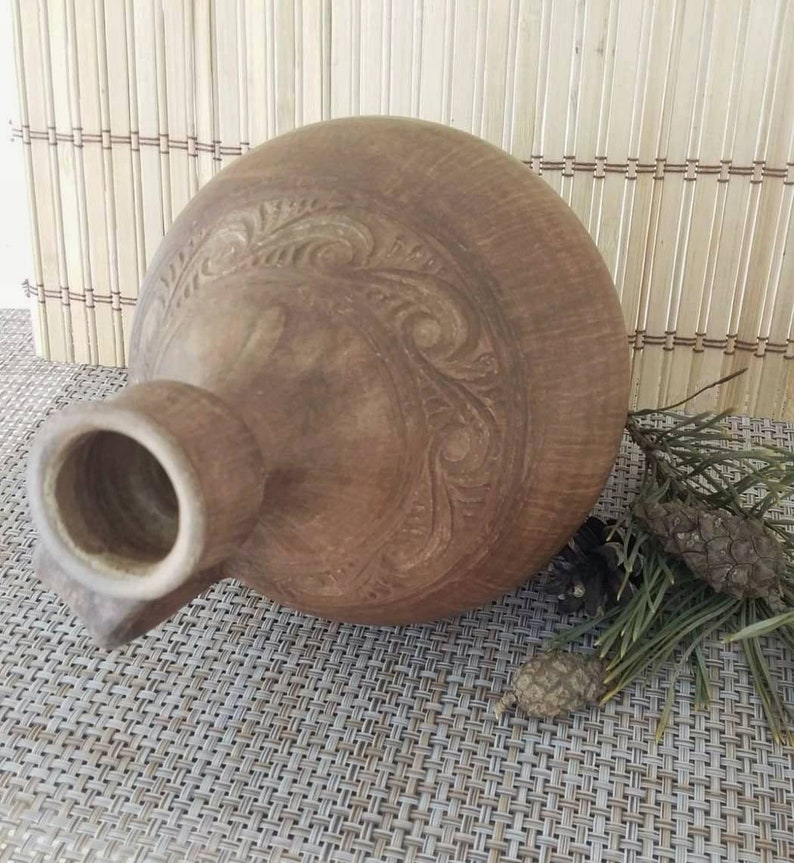 May include: A brown ceramic pitcher with a carved design. The pitcher has a wide mouth and a narrow neck.