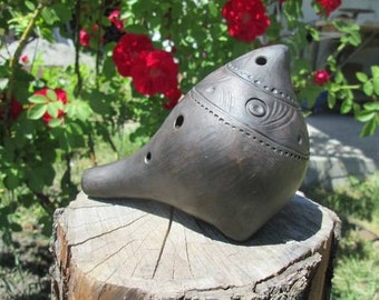 May include: A dark brown clay ocarina shaped like a bird with decorative lines and circles. The ocarina has six finger holes and is sitting on a tree stump.