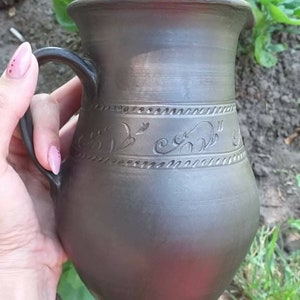 May include: A dark gray ceramic pitcher with a handle and a decorative design of swirling lines and birds. The pitcher is handmade and has a rustic look.