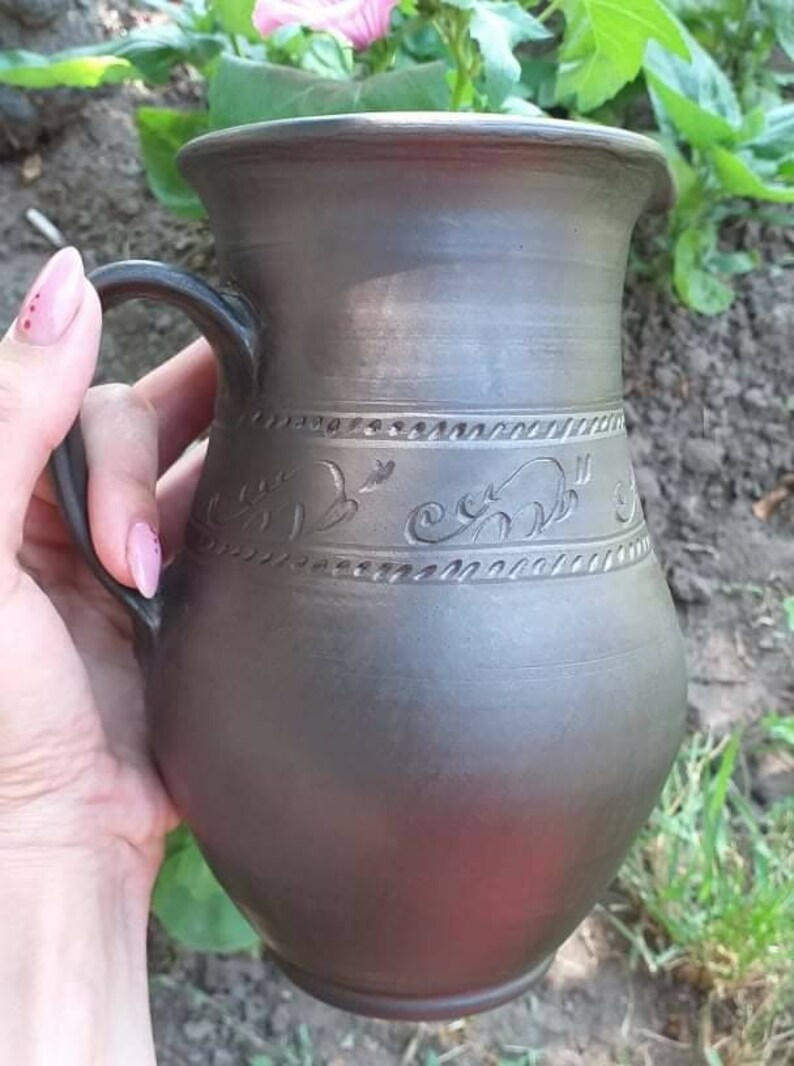 May include: A dark gray ceramic pitcher with a handle and a decorative design of swirling lines and birds. The pitcher is handmade and has a rustic look.