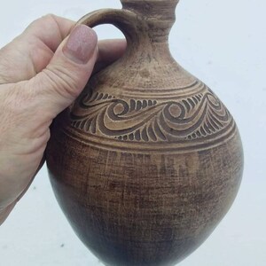 May include: A brown ceramic pitcher with a carved design. The pitcher has a handle and a wide mouth.