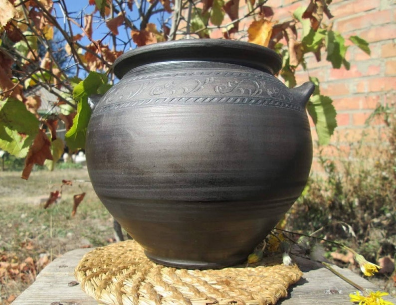 May include: A large, dark brown, ceramic pot with a decorative design around the top. The pot is sitting on a woven straw mat.