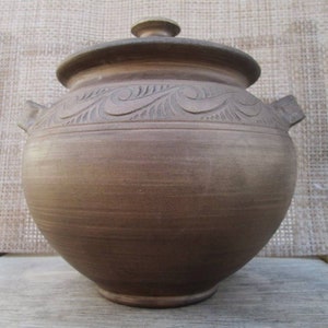 Puede incluir: Una olla de cerámica marrón con tapa y dos asas. La olla tiene un diseño decorativo tallado en el lateral.
