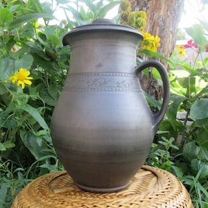 May include: A large, dark brown ceramic pitcher with a lid and a handle. The pitcher has a decorative design around the middle.