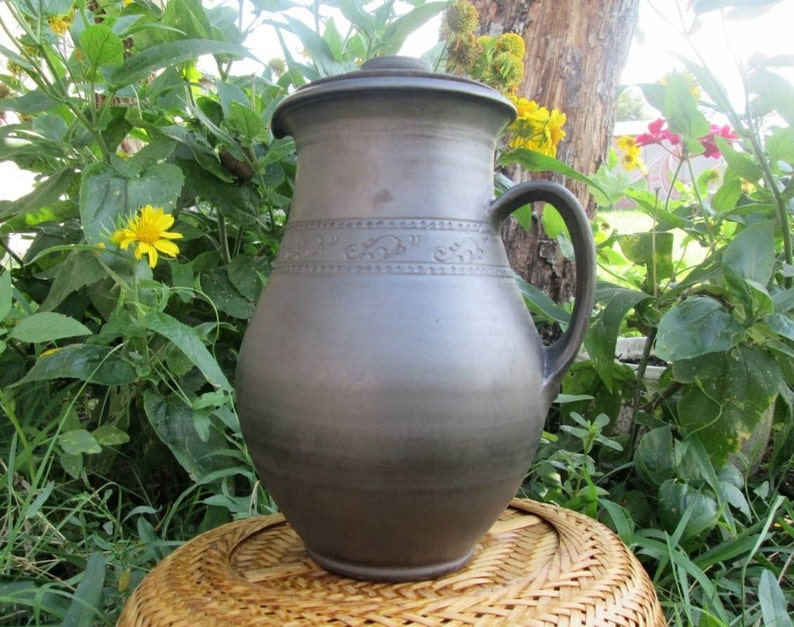 May include: A large, dark brown ceramic pitcher with a lid and a handle. The pitcher has a decorative design around the middle.