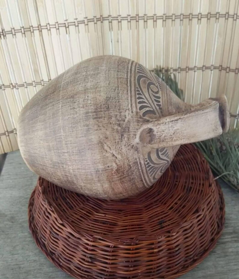 May include: A large, brown, ceramic pitcher with a carved design. The pitcher is sitting on a woven wicker base.