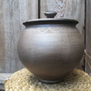 Puede incluir: Una olla de cerámica marrón con tapa, que presenta un sencillo diseño blanco alrededor del borde. La olla está sentada sobre una estera tejida.