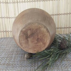 May include: A large, brown, ceramic bowl with a slightly rounded bottom. The bowl is sitting on a woven mat with pine needles and pine cones.