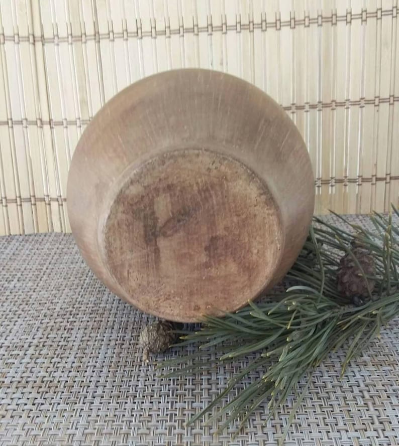 May include: A large, brown, ceramic bowl with a slightly rounded bottom. The bowl is sitting on a woven mat with pine needles and pine cones.