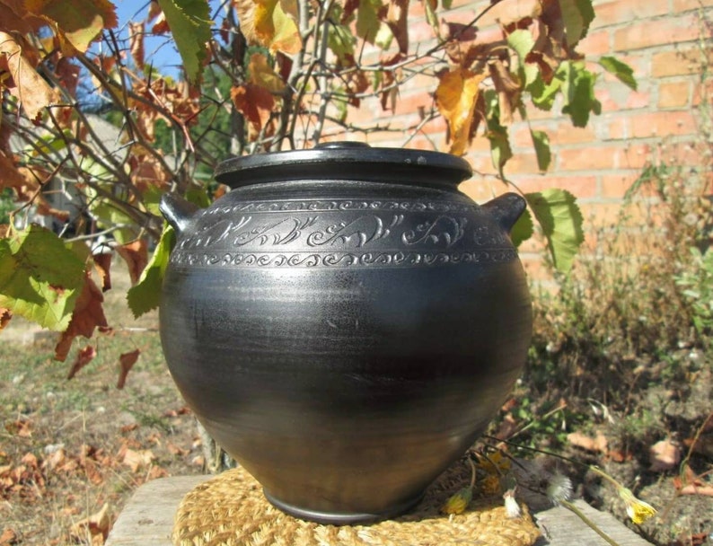 May include: A black ceramic pot with a lid and intricate carvings around the top. The pot is sitting on a woven mat.
