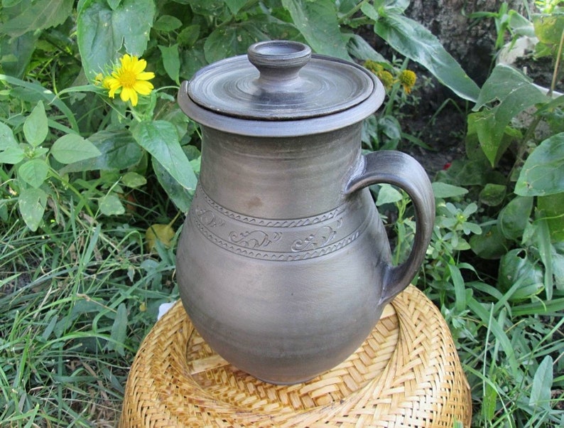 May include: A dark brown ceramic pitcher with a lid and a handle. The pitcher has a decorative design around the middle.