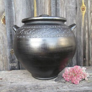 May include: A black ceramic jar with a lid and decorative carvings. The jar is sitting on a wooden surface.