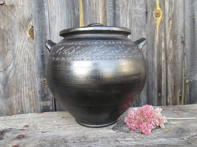 May include: A black ceramic jar with a lid and decorative carvings. The jar is sitting on a wooden surface.